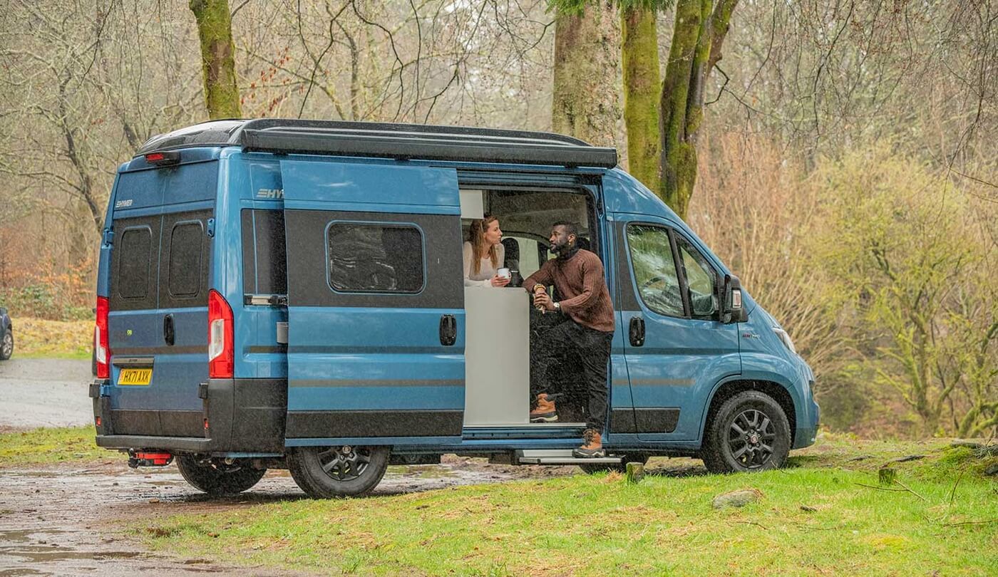 Zwei Personen in der offenen Seitentür eines blauen Campers am Wald.